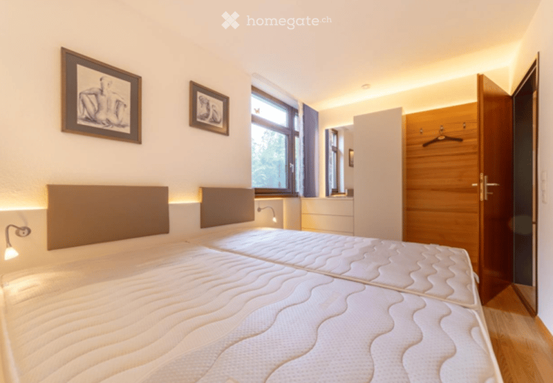 Modern bedroom with two single beds, nightstands, framed artwork, and a wooden wardrobe.