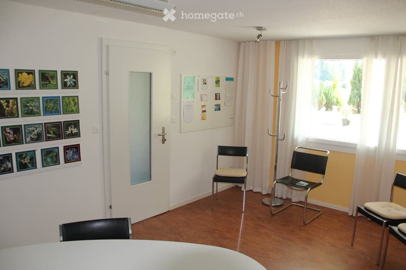 A small, well-lit room with a white table, several black chairs, a coat rack, and a wall adorned with framed pictures. The room has a wooden floor and a large window with white curtains.