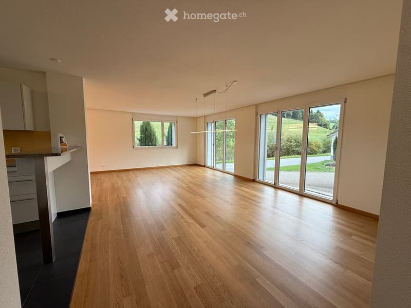 Bright, empty room with wooden flooring, large windows, and sliding glass doors offering a scenic view of the countryside.