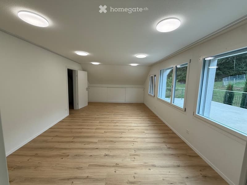 Empty room with wooden flooring, white walls, and large windows offering a view of the outdoors. The room features recessed lighting and an open doorway leading to another space.