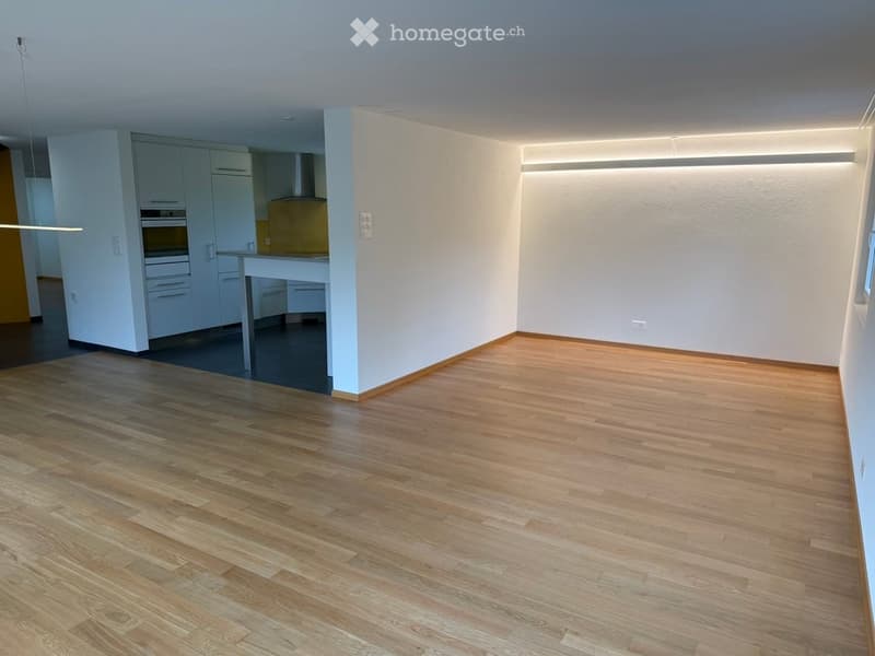Empty room with wooden flooring, white walls, and a modern kitchen area visible in the background.