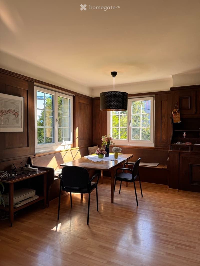 Brightly lit room with wooden paneling, hardwood floor, large windows, a dining table with chairs, a sofa, and a laptop on a side table.