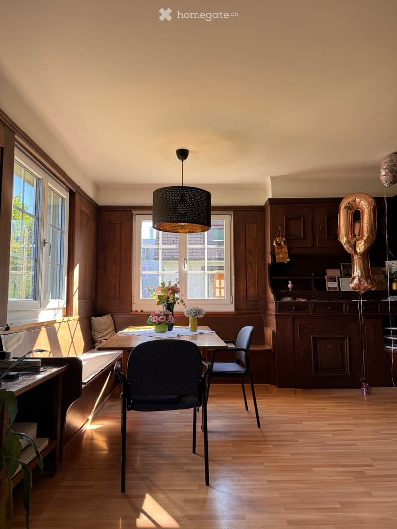 Brightly lit dining area with wooden floors, large windows, a round wooden table with chairs, a pendant light fixture, and a cabinet with decorative items and balloons.