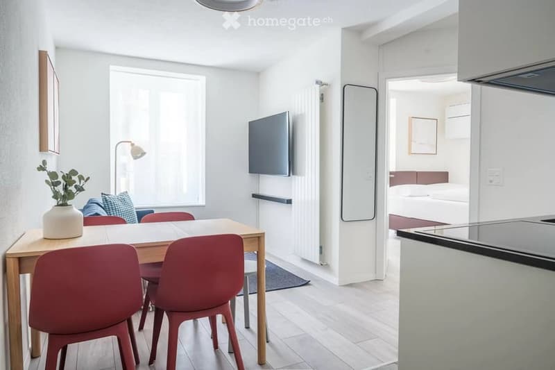 A modern, open-concept living and dining area with a wooden dining table, red chairs, a sofa with pillows, a floor lamp, a television, a mirror, and a bed visible in the background. The space is bright with large windows and neutral tones.