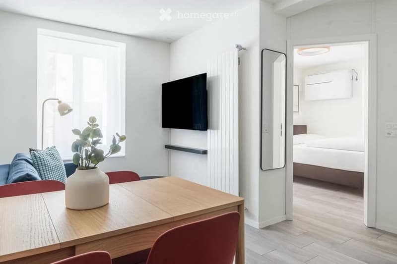 A modern, well-lit room with a wooden table, chairs, a sofa with pillows, a vase with a plant, a television mounted on the wall, and a mirror. An adjacent bedroom with a bed is visible through an open doorway.