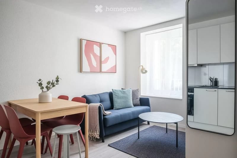 Modern, open-concept living and dining area with a blue sofa, wooden dining table, red chairs, and a small kitchen area visible in the background.