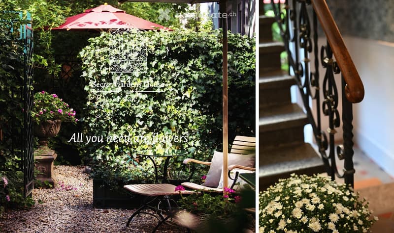 A serene garden area with a wooden bench, glass table, hanging basket, umbrella, and lush greenery, including ivy-covered walls and flowering plants.