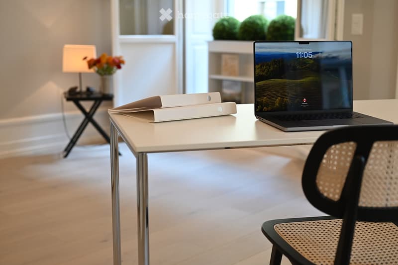 A modern, well-lit room with a white table holding a laptop and a small device, a wicker chair, a side table with a lamp and flowers, and a large window with a view of greenery.