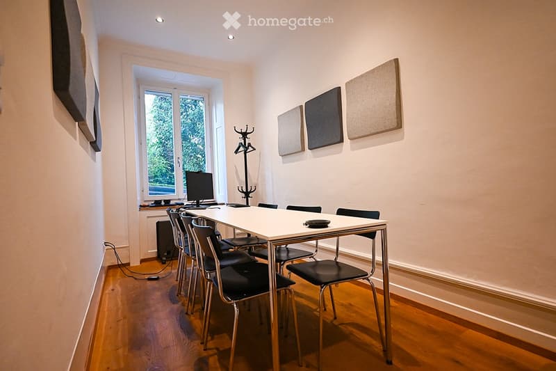 A small, modern dining or study area with a white table, black chairs, a computer, and a coat rack near a large window.