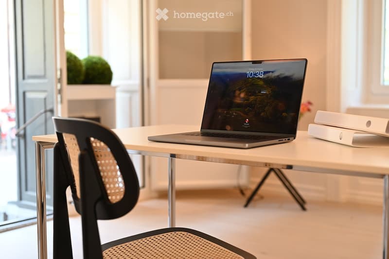 A modern, well-lit room with a white table, a laptop displaying a scenic wallpaper, a black ergonomic chair, and a stack of books on the right side.