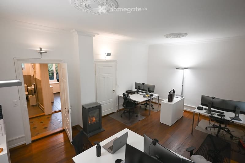 Bright, modern office space with multiple desks, chairs, laptops, monitors, and a small fireplace. The room features wooden flooring and white walls.