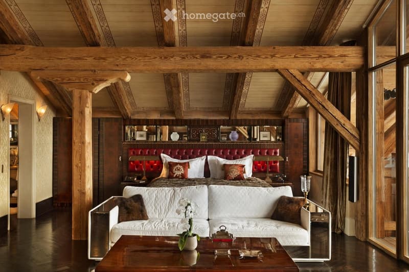 Luxurious bedroom with exposed wooden beams, a large red leather headboard bed, a white sofa, a coffee table with a flower vase, and wall-mounted lamps.