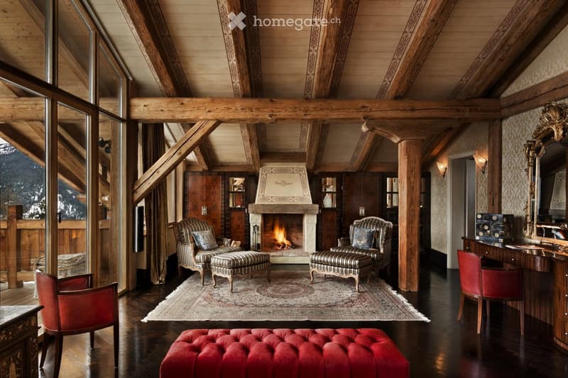 Luxurious living room with exposed wooden beams, a stone fireplace, patterned armchairs, a red velvet ottoman, and a large mirror. The room features a wooden desk and a patterned rug.