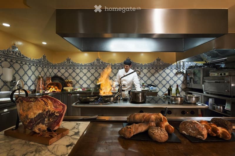 A chef is cooking in a professional kitchen with a large stove, pots, pans, and a variety of food items including bread and cured meat. The kitchen features a decorative tile backsplash and a large exhaust hood.