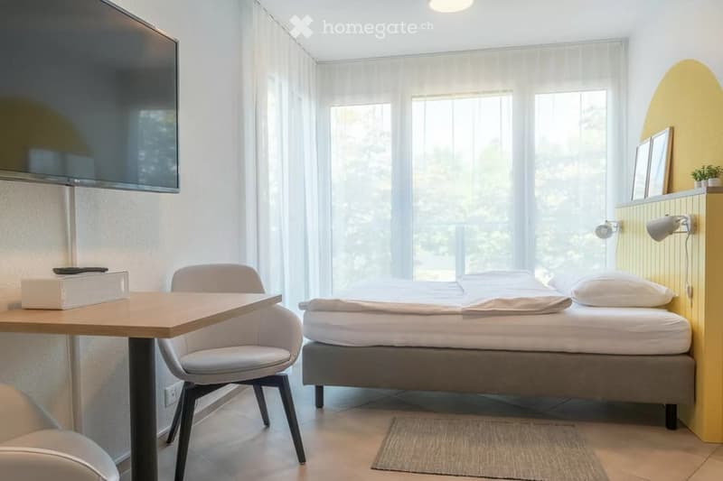 Modern bedroom with a double bed, a small table with a chair, a wall-mounted TV, and a yellow accent wall with framed pictures.