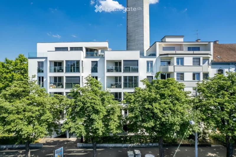 Modern multi-story residential building with white facades, large windows, and multiple balconies. Surrounded by lush green trees.