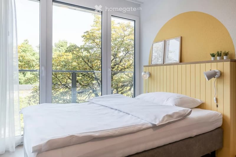 Bright bedroom with a large window and balcony access, featuring a neatly made bed, nightstand with lamp, and decorative picture frames on the wall.