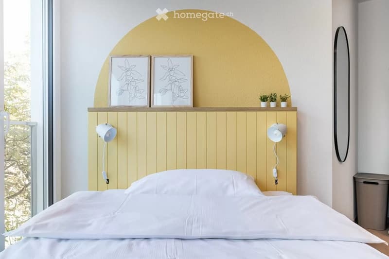 Modern bedroom with a white bed, yellow accent wall, wall-mounted lamps, and decorative picture frames above the bed.
