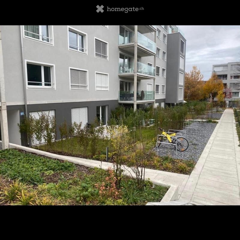 Modern multi-story apartment building with balconies, surrounded by landscaped greenery and a paved walkway.