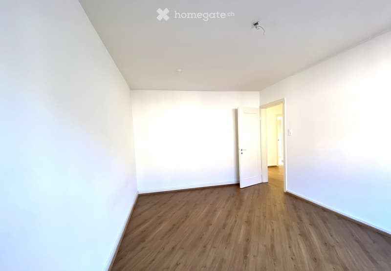 Empty room with white walls and wooden flooring, featuring a partially open door leading to another room.