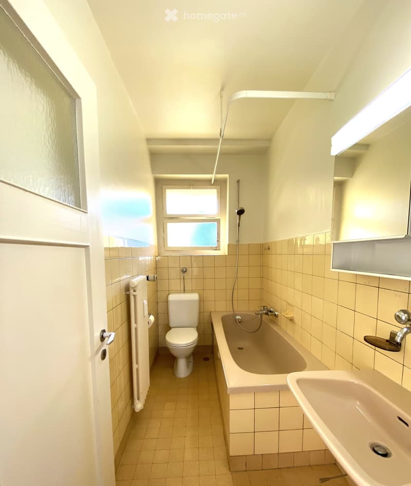 Bathroom with a toilet, bathtub, sink, radiator, and a window with frosted glass panes. The walls are tiled in beige, and the floor is tiled in light brown.