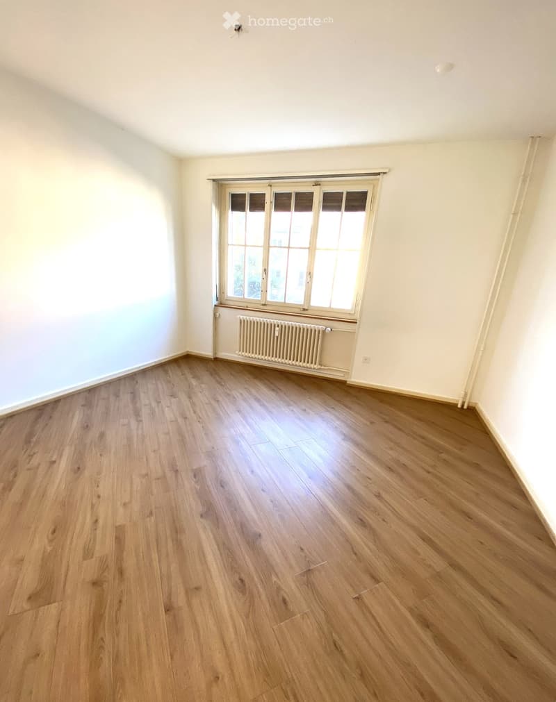 Empty room with wooden flooring, white walls, and a large window with blinds. A radiator is visible below the window.