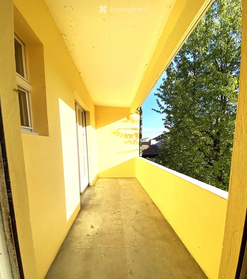 A narrow balcony with yellow walls, a concrete floor, and a view of greenery outside.