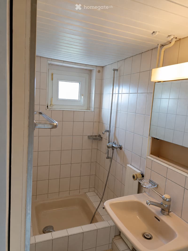 A compact bathroom with a bathtub, sink, mirror, and showerhead. The walls and floor are tiled, and there is a small window above the bathtub.