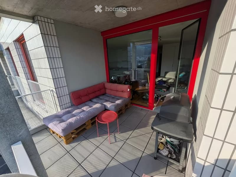 Balcony with a modern design, featuring a grey sectional sofa, a small pink table, and a grey barbecue. The space includes a tiled floor and a view of the city.