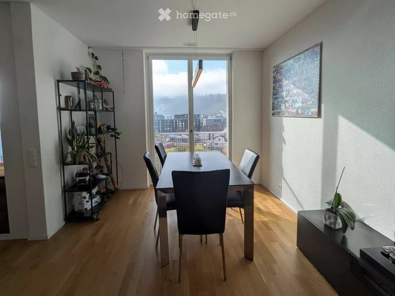 Bright dining area with wooden flooring, large window offering a view, dining table with chairs, shelf with plants and items, and a picture frame on the wall.