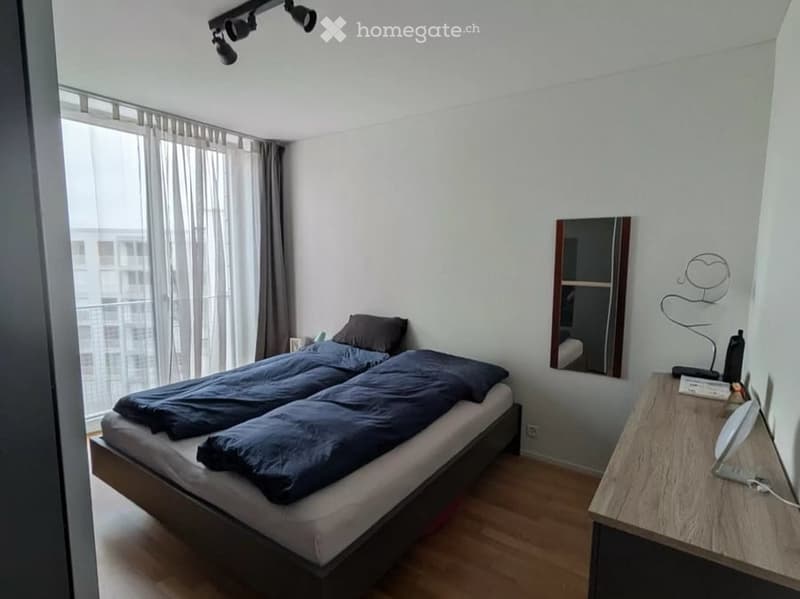 Modern bedroom with a double bed, nightstand, mirror, and large window with curtains. The room features wooden flooring and track lighting.