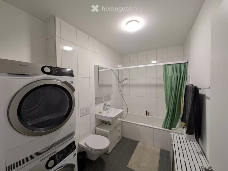 Modern bathroom with white tiled walls, a sink, toilet, bathtub with shower, mirror, washing machine, dryer, and a green shower curtain.