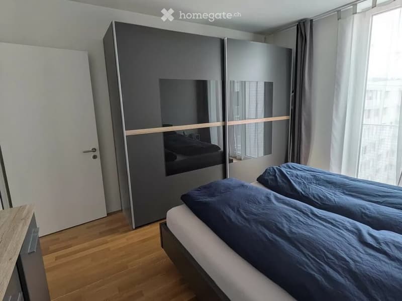 Modern bedroom with a large mirrored wardrobe, a neatly made bed with dark blue bedding, and wooden flooring.