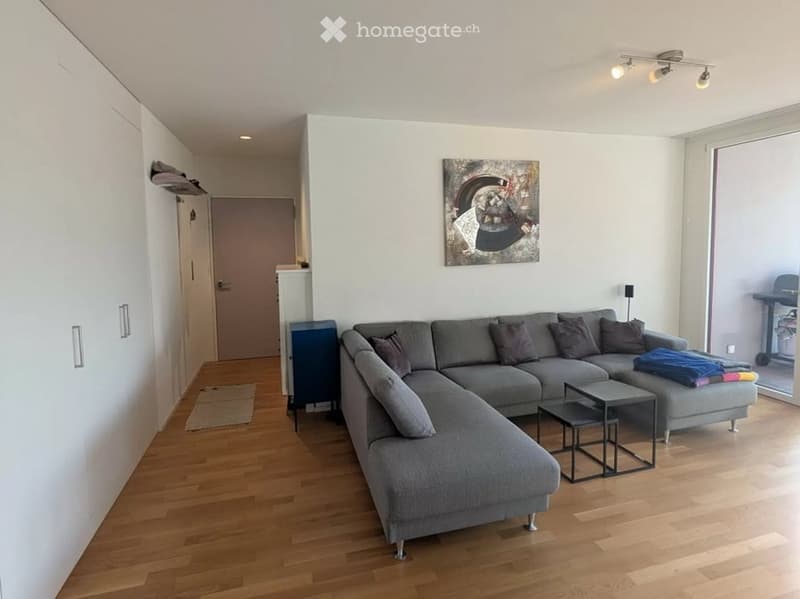 Modern living room with a grey sectional sofa, coffee table, side table, and an art piece on the wall. The room features wooden flooring and built-in storage units.
