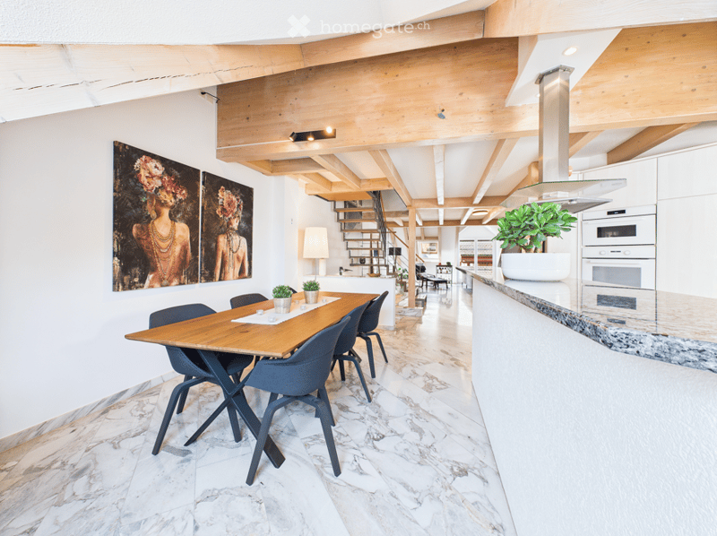 Open-plan dining and kitchen area with a wooden dining table, modern chairs, marble flooring, and white kitchen cabinets. A staircase leads to an upper level, and artwork adorns the walls.