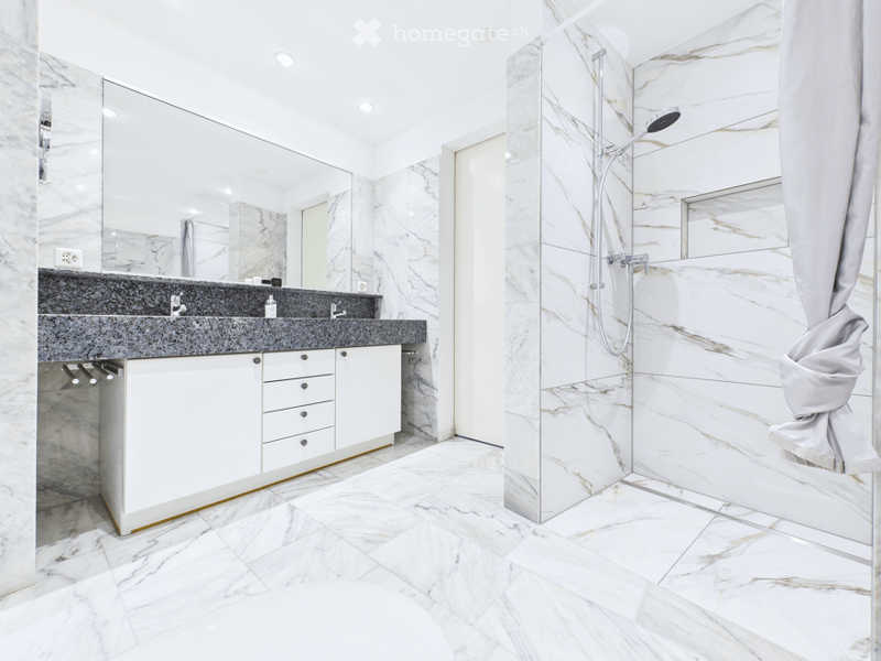 Modern bathroom with marble tiles, double sink, large mirror, and a walk-in shower with glass enclosure.