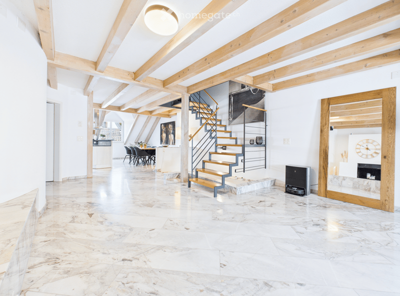 Bright, open hallway with exposed wooden beams, marble flooring, and a modern staircase leading to an upper level. The space features a dining area with chairs and a table visible in the background.