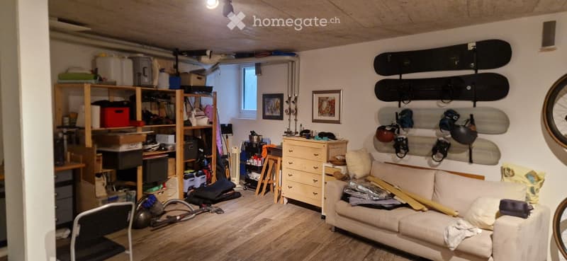 A cozy, multi-purpose room with a sofa, a chair, a shelf filled with storage boxes and items, a small table, a dresser, and several snowboards mounted on the wall. The room also features a laptop on the floor and a vacuum cleaner nearby.