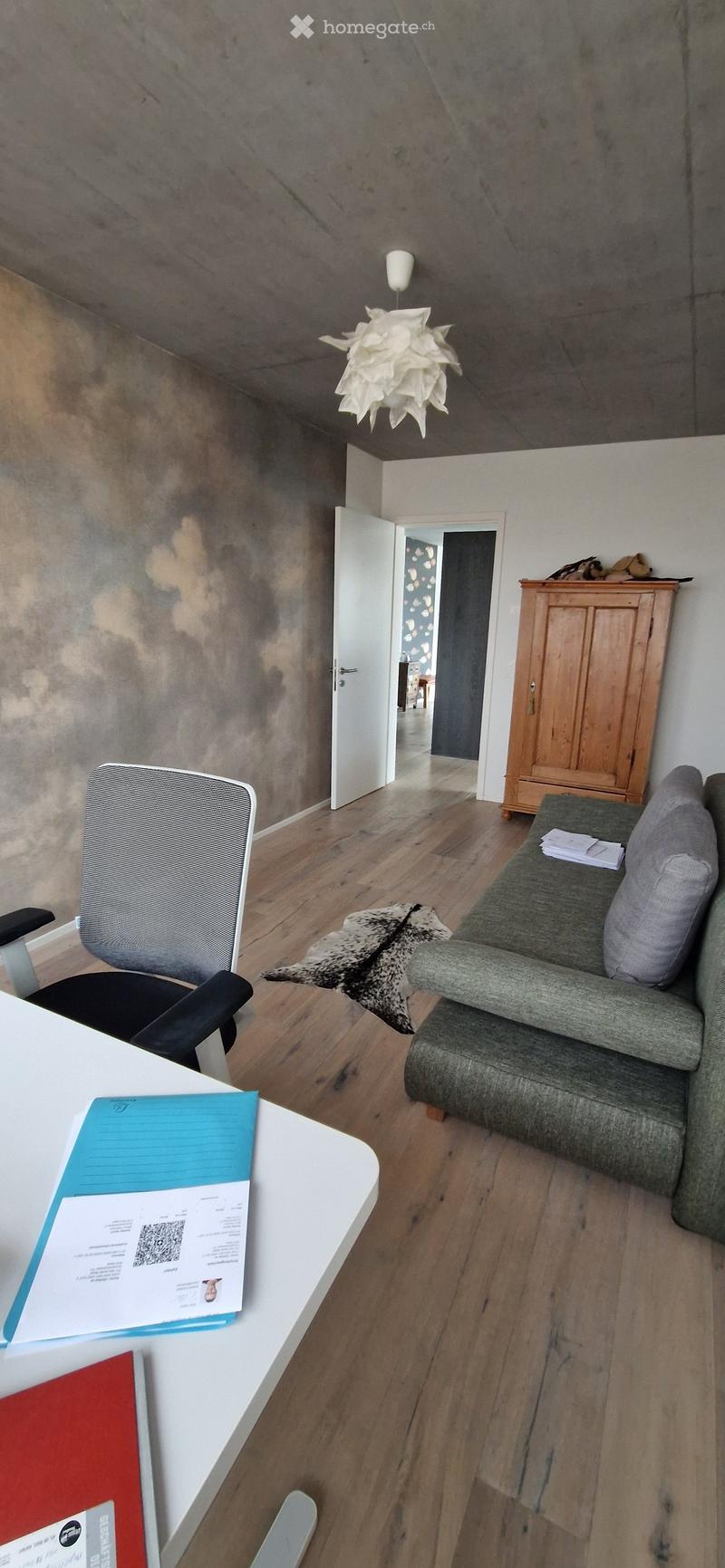 A modern living room with a grey sofa, a wooden cabinet, a hanging lamp, a desk with papers, and a chair.