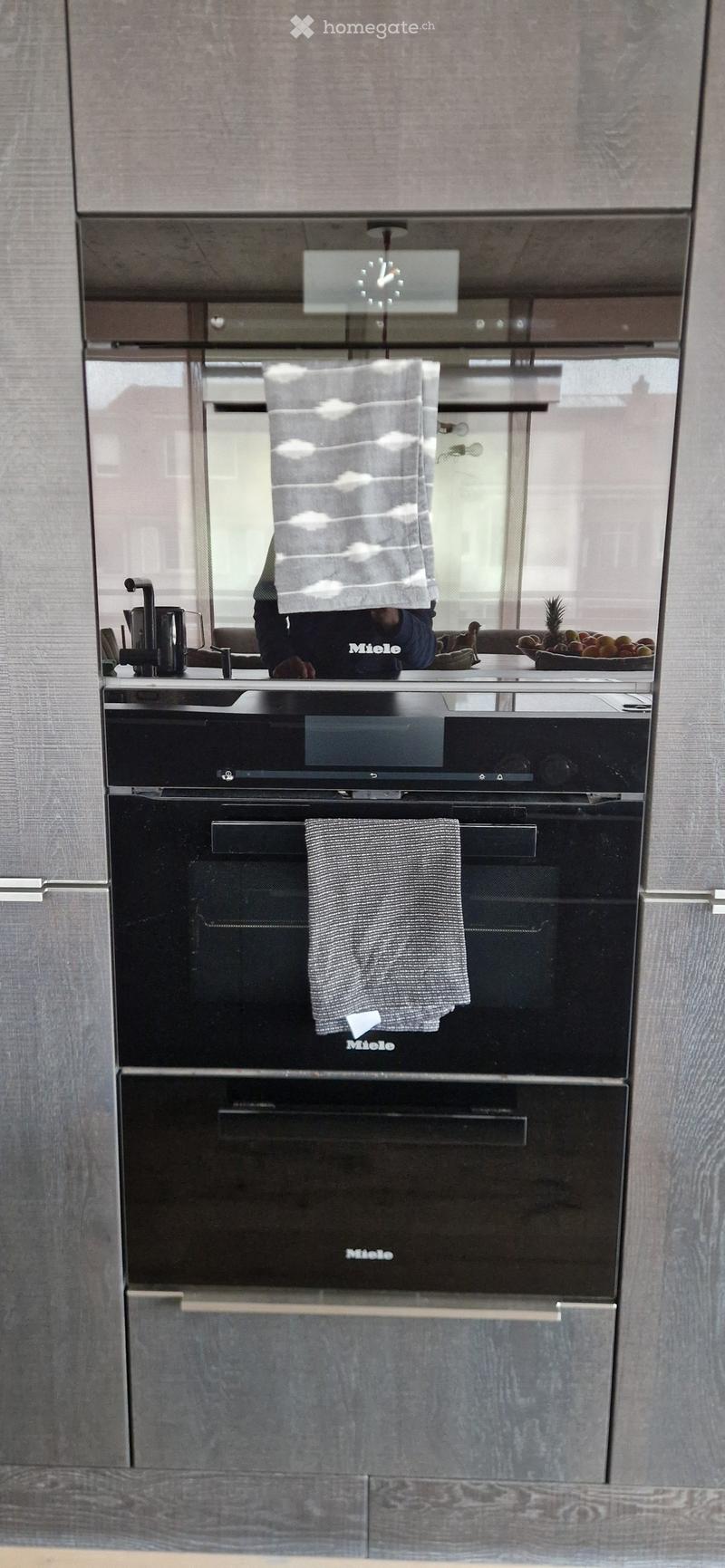 Modern kitchen with stainless steel oven, towels hanging on the oven door, coffee maker on the counter, and a bowl of fruit in the background.