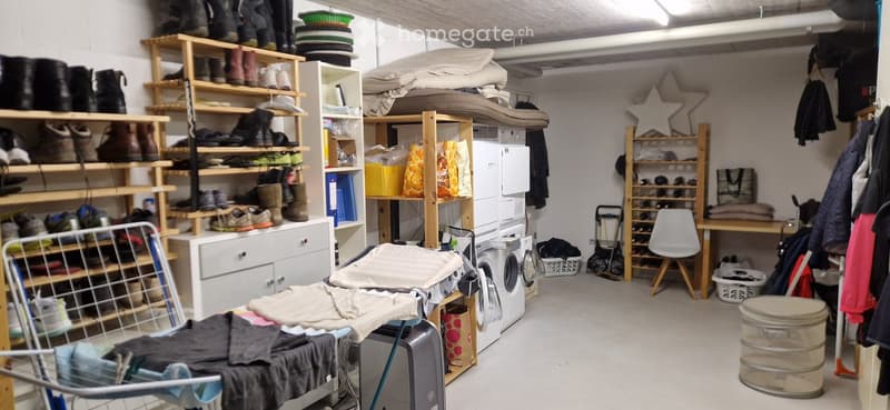 A well-organized storage room with multiple shelves filled with shoes, clothing, and other items. A washing machine and refrigerator are visible, along with a chair, table, and various storage bins.