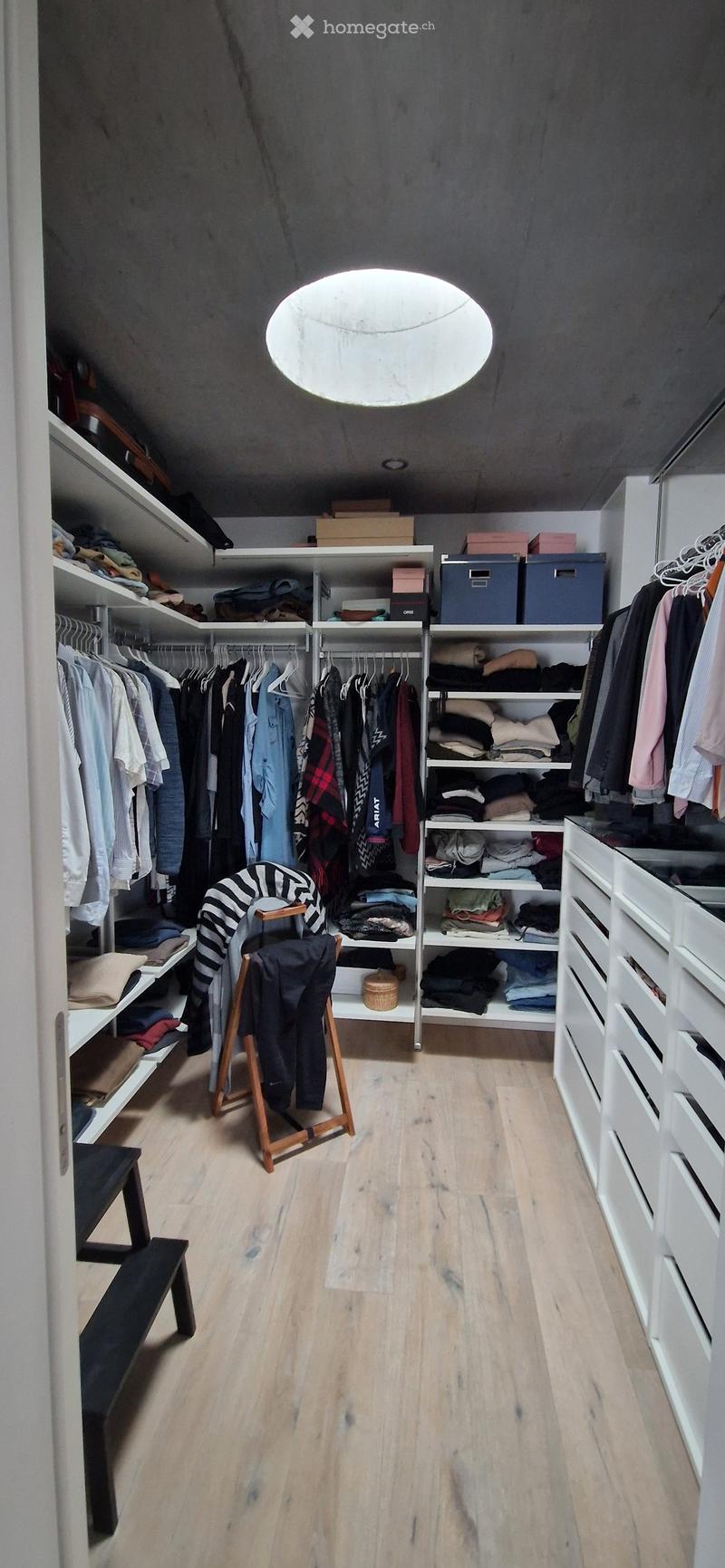 Well-organized walk-in closet with wooden flooring, white built-in wardrobes, shelves, and storage boxes. A small wooden stool and bench are placed in the center.