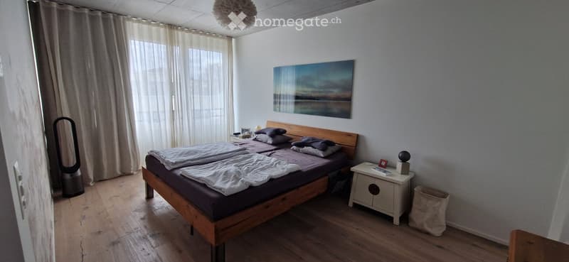 A well-lit bedroom with a wooden bed, nightstand, and a large window with sheer curtains. The room features a painting on the wall and a fan near the door.