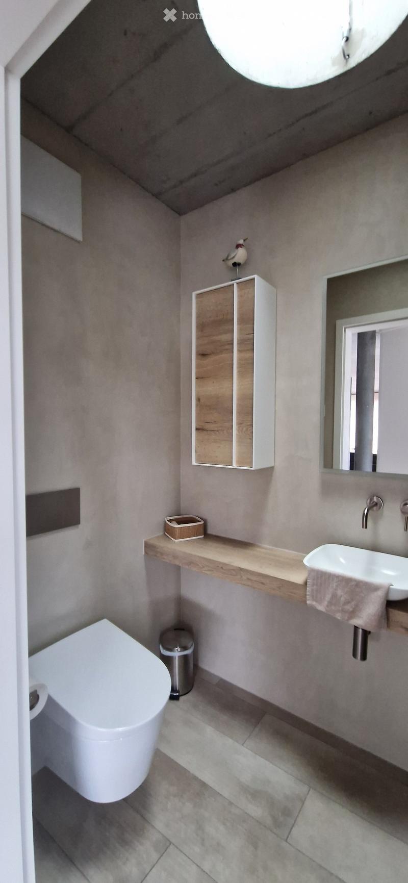 Modern bathroom with a white toilet, floating wooden shelf, sink, mirror, and decorative bird on a cabinet.