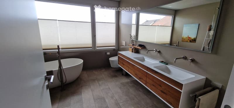Modern bathroom with a freestanding bathtub, double sink vanity with wooden cabinets, large mirror, and a toilet. The space features large windows with blinds and tiled flooring.