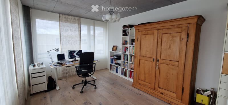 A modern, well-lit study or office space with a wooden floor, large windows with sheer curtains, a white desk with a laptop and monitor, a black office chair, a tall wooden cabinet, and a bookshelf filled with books and decorative items.