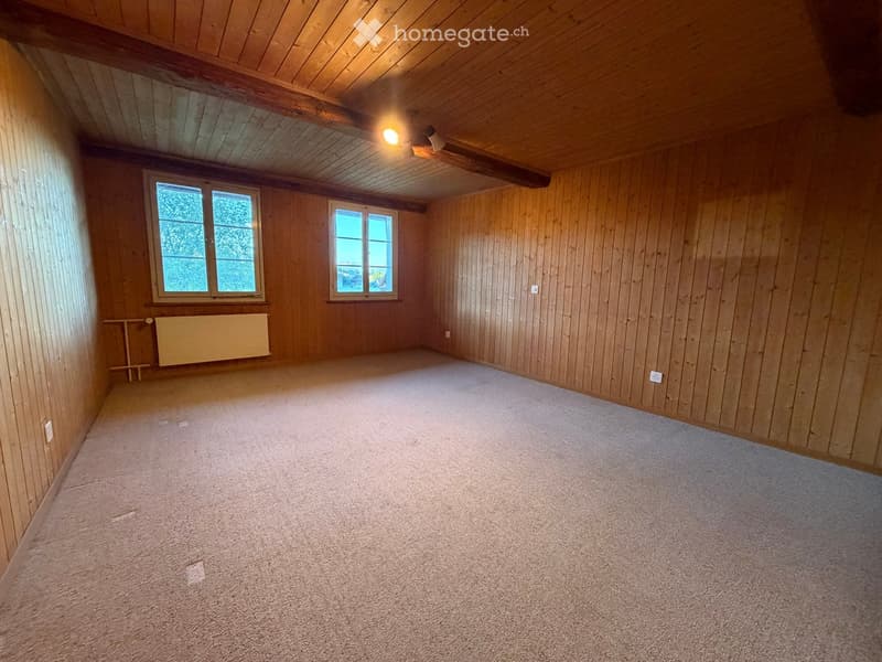 Empty room with wooden walls, carpeted floor, and two windows. Features a ceiling fan and a radiator on one wall.