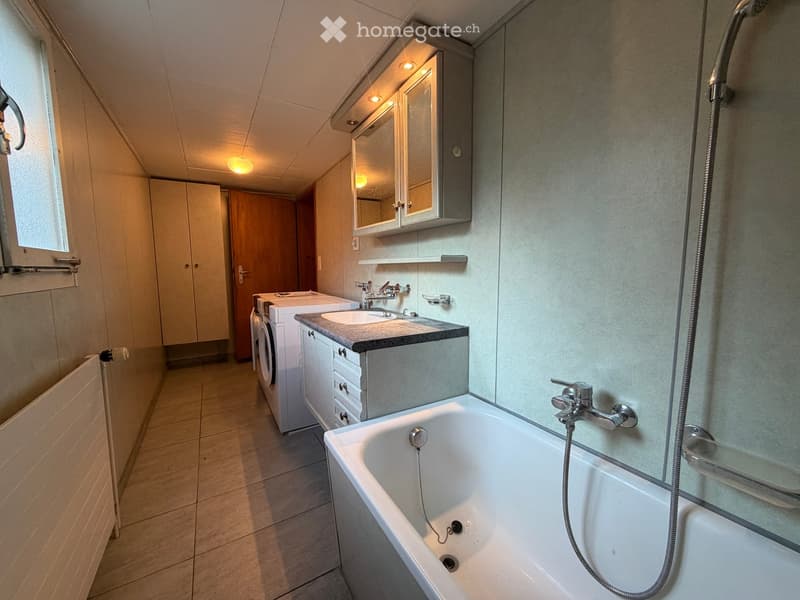 Bathroom with a bathtub, sink, mirror, and a private washing machine. The space features tiled floors and walls, with a window providing natural light.