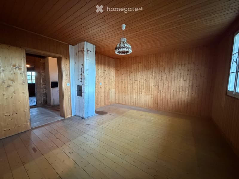 Empty room with wooden walls, wooden floor, and a hanging light fixture. There is an open doorway leading to another room.