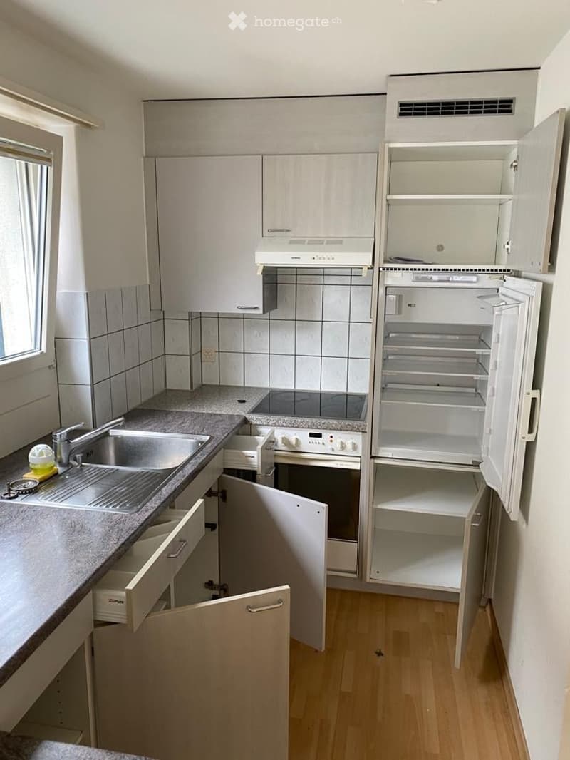 Modern kitchen with light-colored cabinets, a stainless steel sink, an oven, and an open refrigerator. The countertop has a few small items, and the floor is wooden.